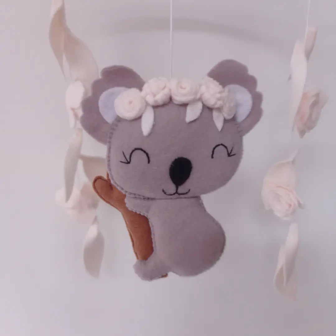 Koala gris en feutrine sur branche d'arbre avec couronne de fleurs blanches.